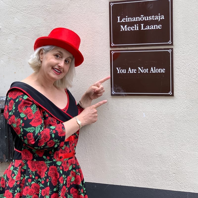 Christine Filip, wearing a red/black floral dress and red hat, points to signs on the wall with text in English and Estonian. The text written on the signs means: You are not alone.